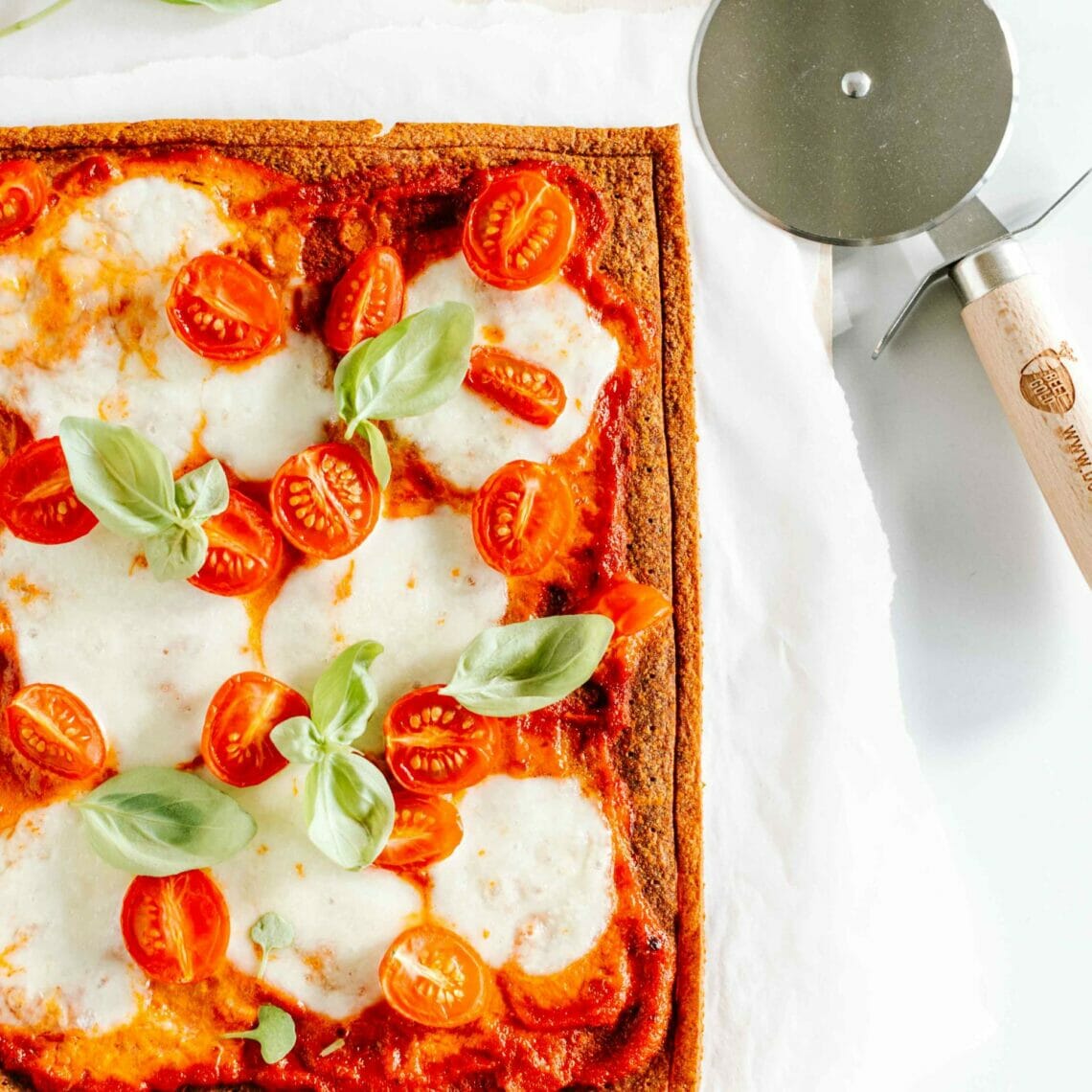 Pizzroller neben einer fertigen Pizza Margherita, belegt mit Tomatensauce, Cocktailtomaten, Mozzarella und Basilikum