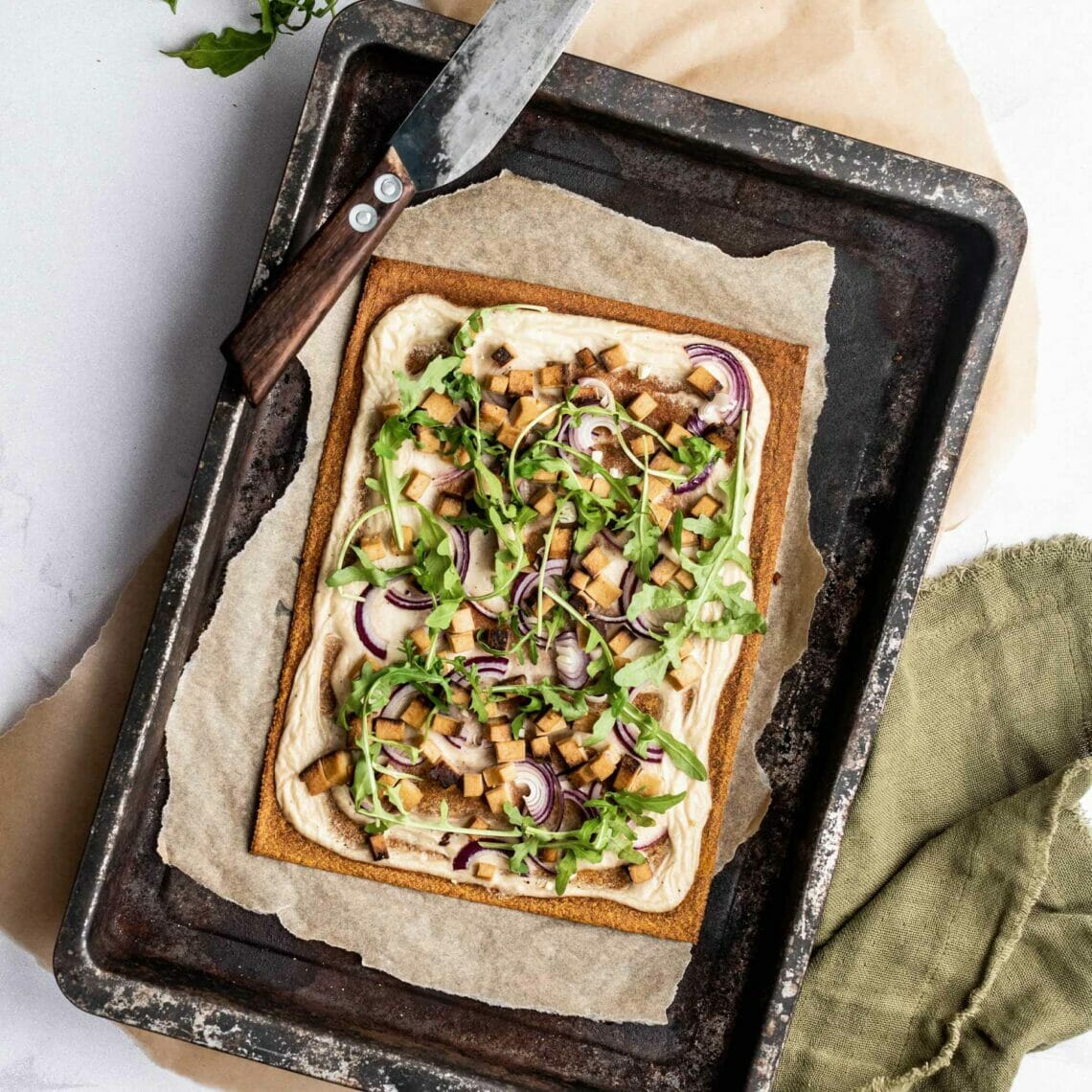 Gemüse Pizzaboden verwendet als Flammkuchen auf einem mit Backpapier belegtem Backblech. Der Flammkuchen ist mit Rucola, Zwiebeln und Räuchertofu belegt