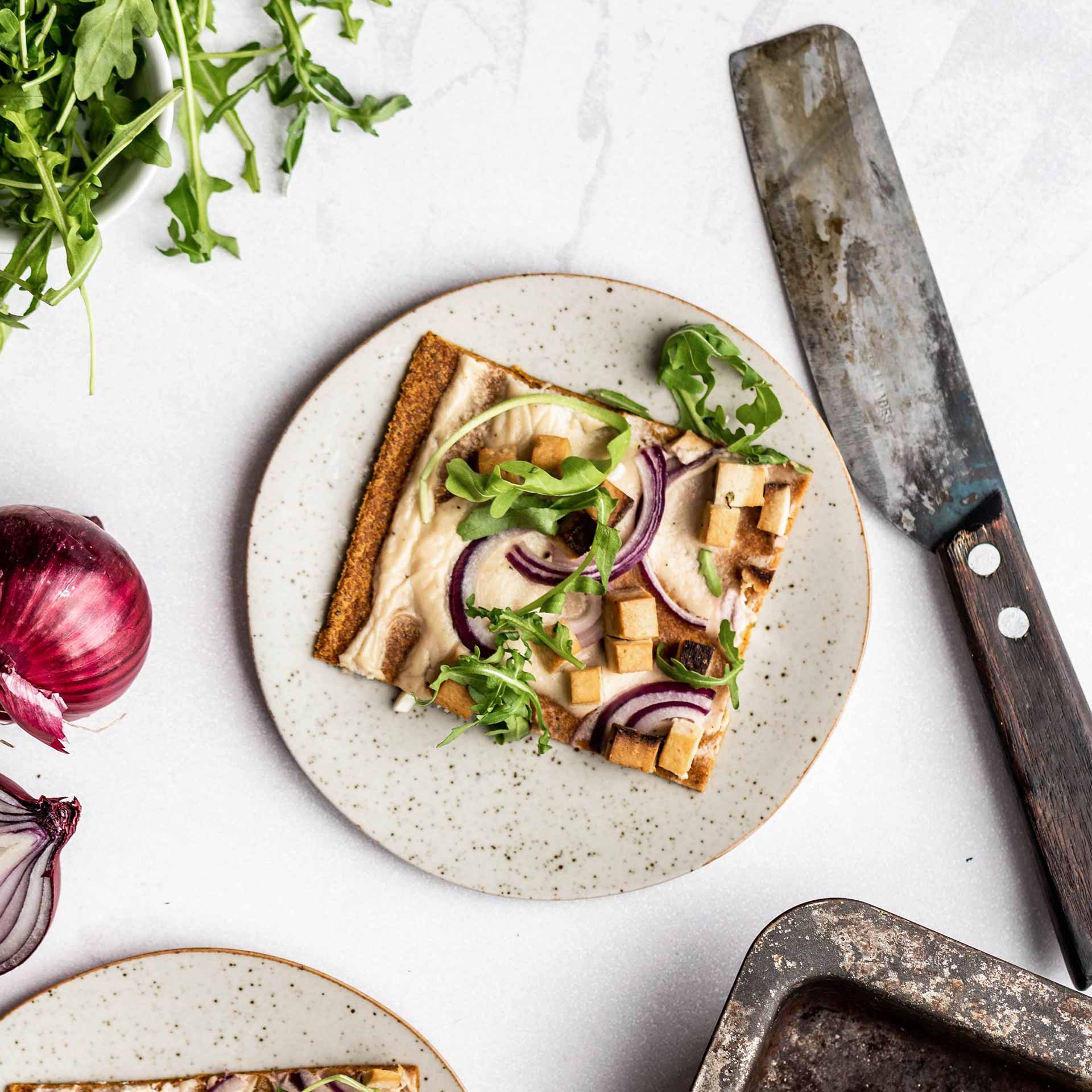 Ein Stück Flammkuchen aus dem Gemüse Pizzaboden belegt mit Frischkäse, Räuchertofu, Roten Zwiebeln und Rucola