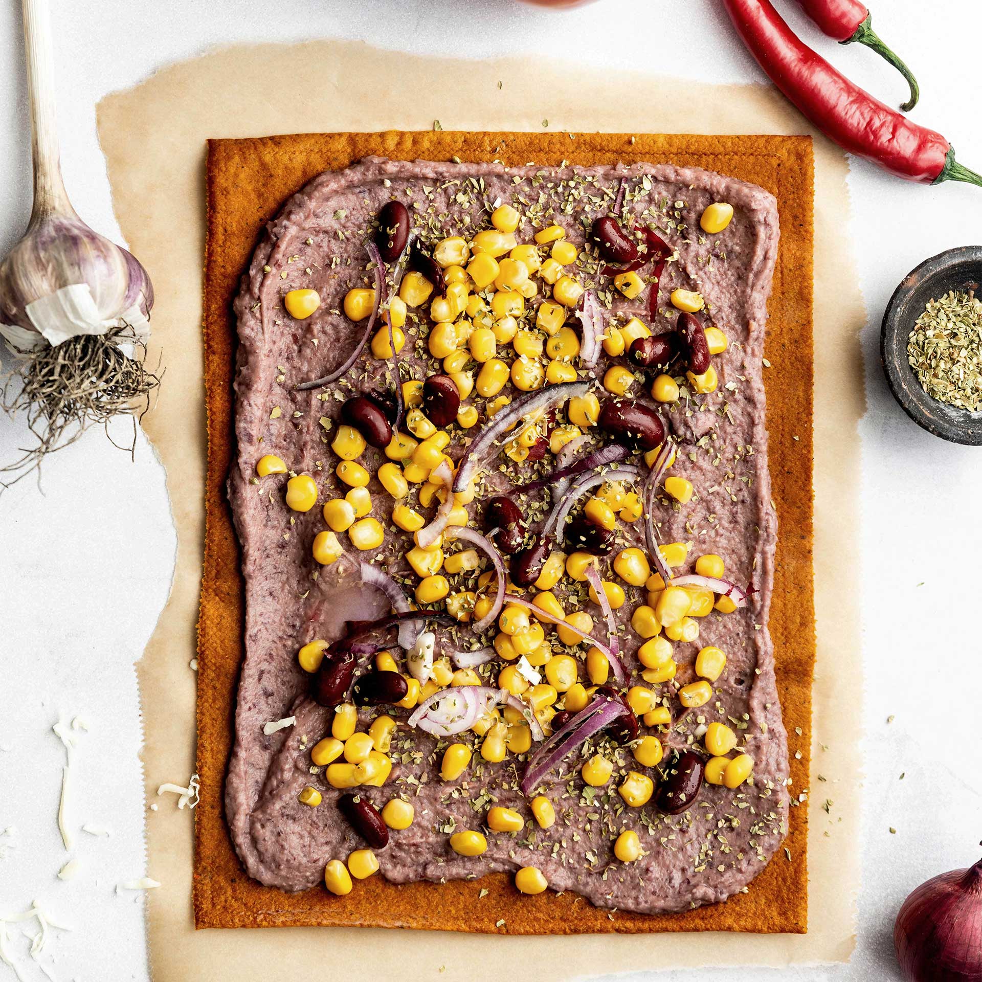 Ungebackener Beetgold Pizzaboden auf Backpapier belegt mir Bohnenmus, Mais, Kidneybohnen und Zwiebeln