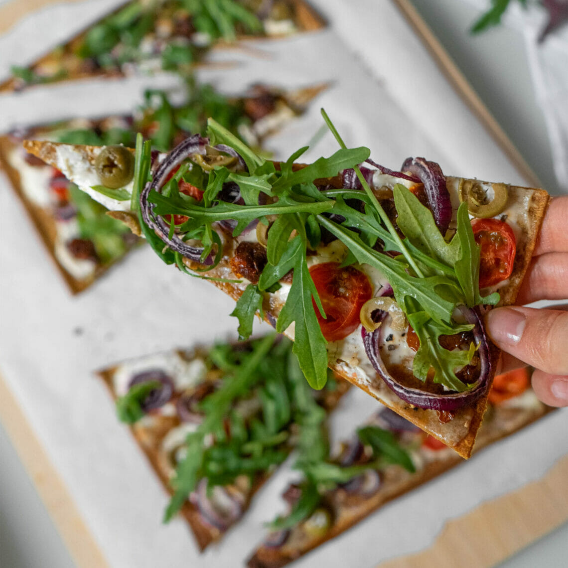 In Hand gehaltenes Flammkuchenstück belegt mit Creme Fraîche belegt mit Tomaten, getrockneten Tomaten, Oliven, roten Zwiebeln und Rucola