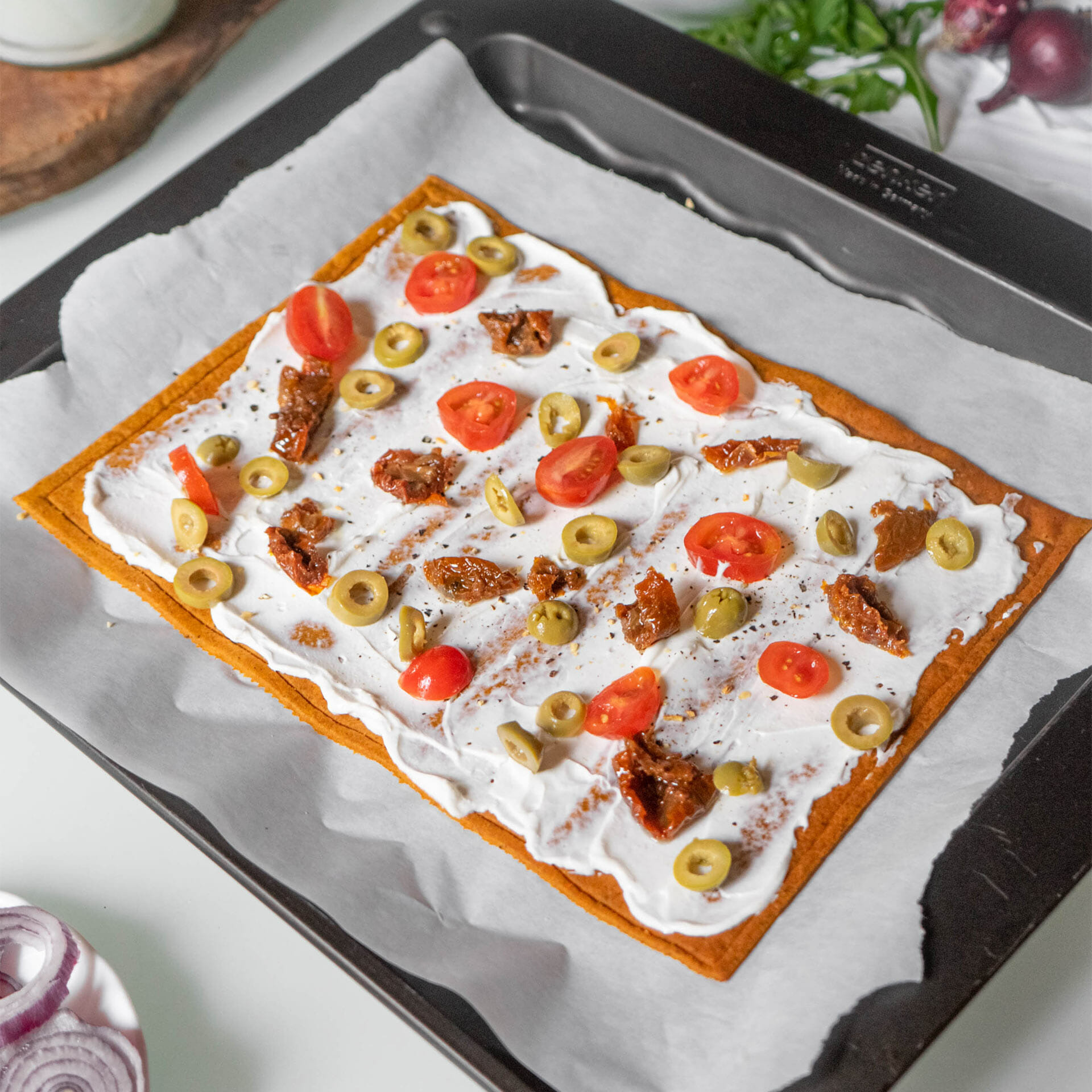 ungebackener Pizzaboden als Flammkuchen mit Creme Fraîche belegt mit Tomaten, getrockneten Tomaten und Oliven