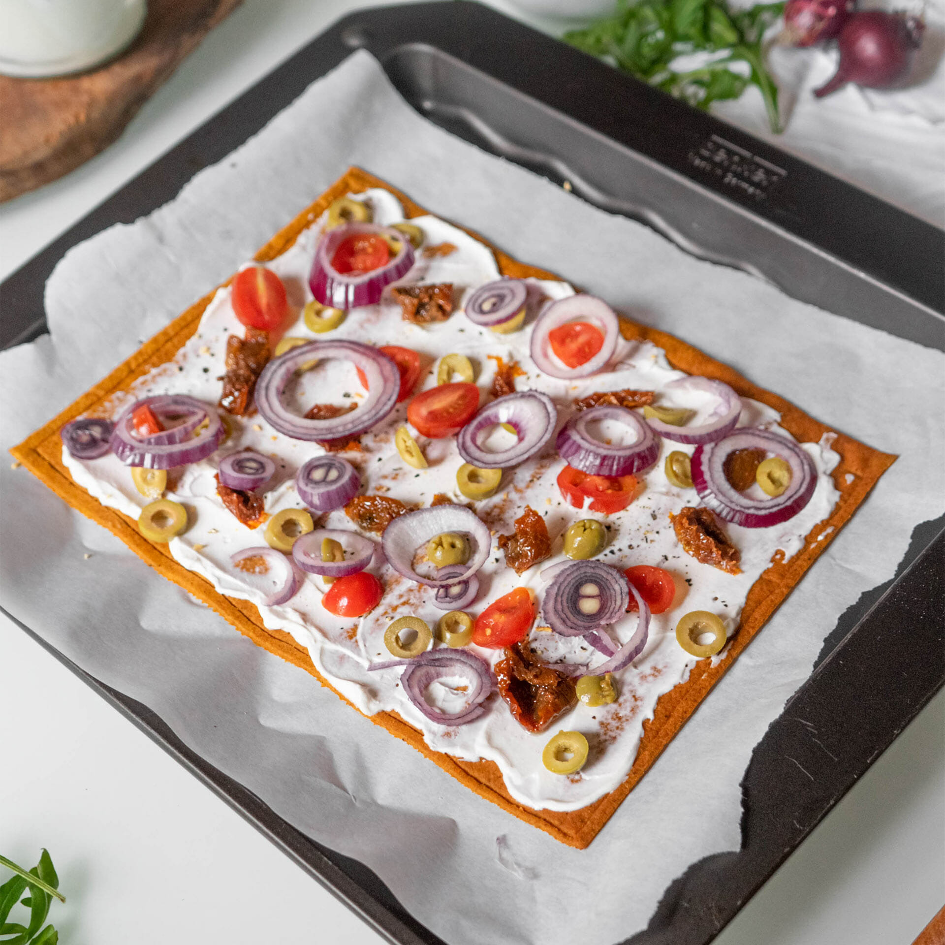Pizzaboden als Flammkuchen mit Creme Fraîche belegt mit Tomaten, getrockneten Tomaten, Oliven, roten Zwiebeln noch ungebacken