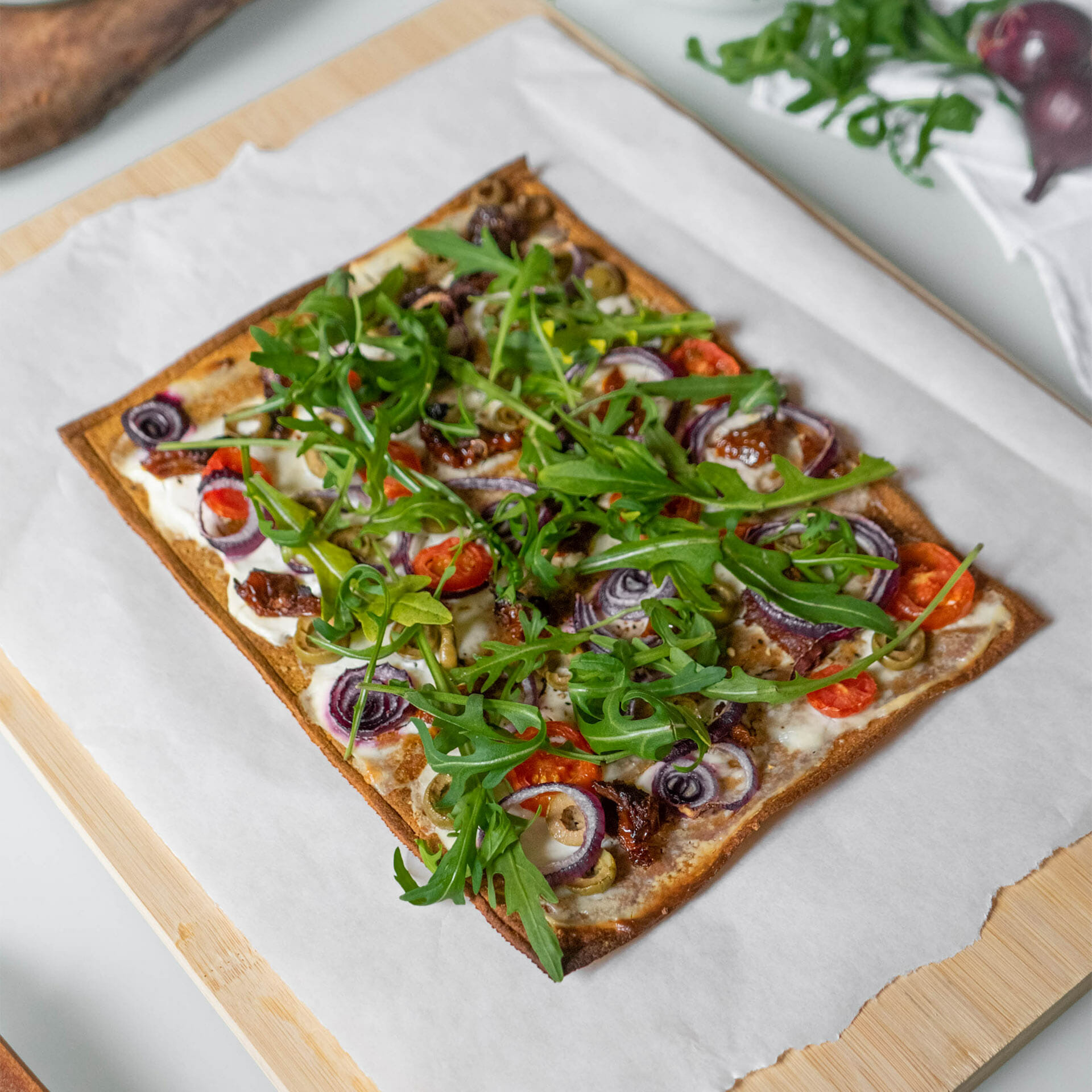 Pizzaboden als Flammkuchen mit Creme Fraîche belegt mit Tomaten, getrockneten Tomaten, Oliven, roten Zwiebeln und Rucola