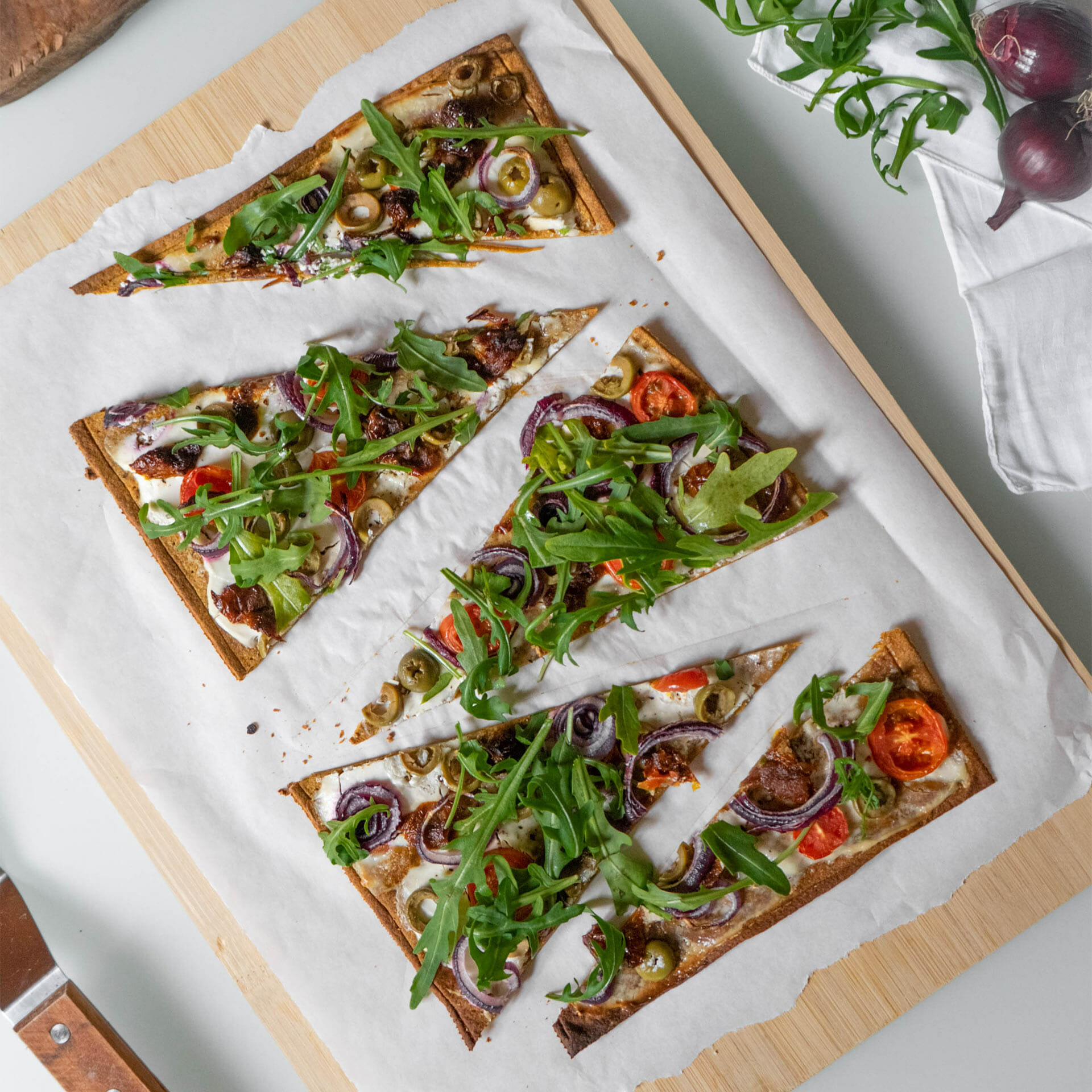 In Dreiecke geschnittener Pizzaboden als Flammkuchen mit Creme Fraîche belegt mit Tomaten, getrockneten Tomaten, Oliven, roten Zwiebeln und Rucola