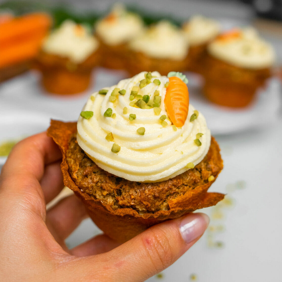 In Hand gehaltener Rübli Muffin mit Buttercreme, Pistazien und Marzipankarotte in Karotten Tortilla Muffinform