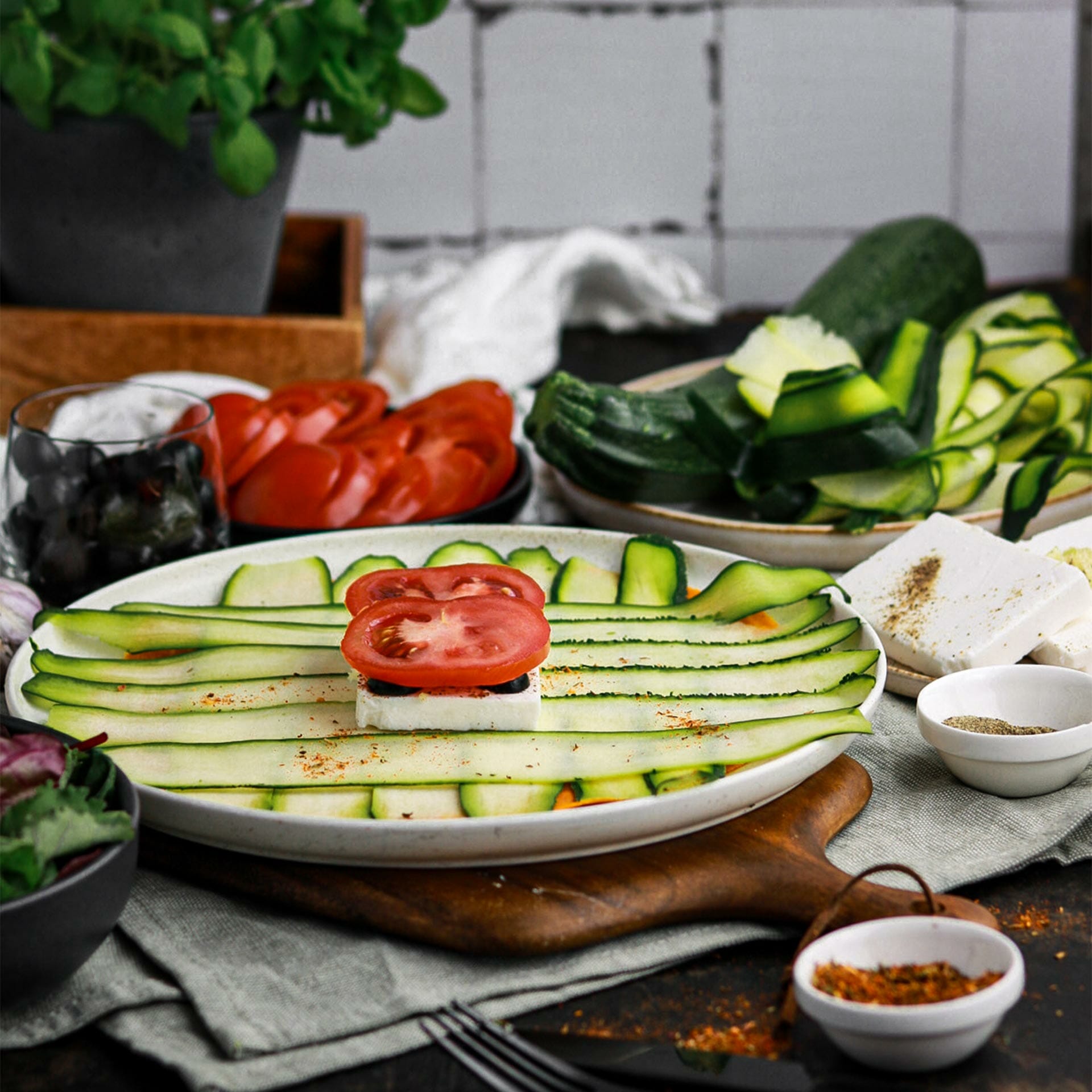 Teller mit Karotten Tortilla belegt mit dünnen Zucchini Streifen, darauf ein Feta Block mit Oliven und Tomatenscheiben