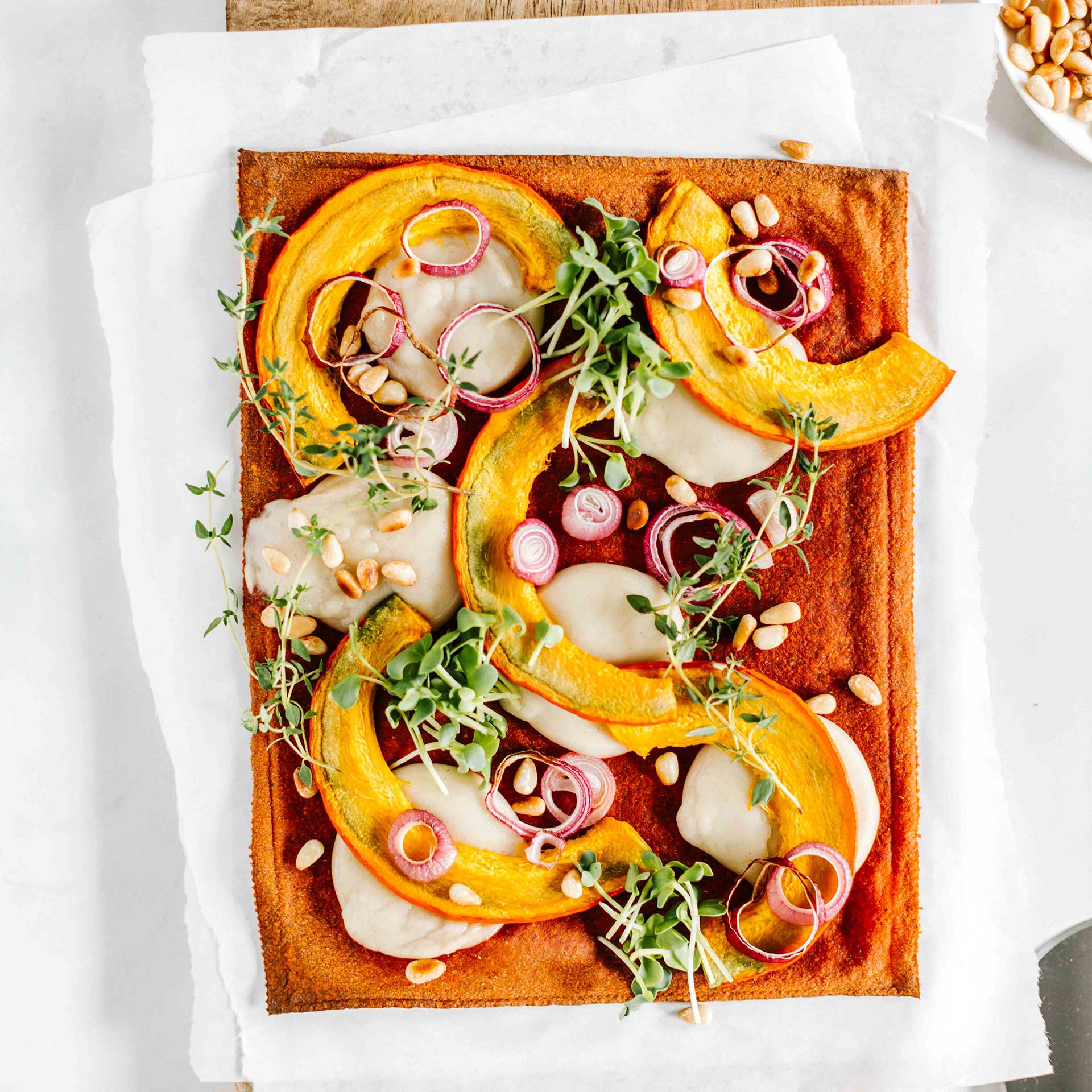 Gebackener Pizzaboden auf Holzbrett belegt mit Mozzarella, Kürbis, Zwiebeln, Pinienkernen, Sprossen und Thymian