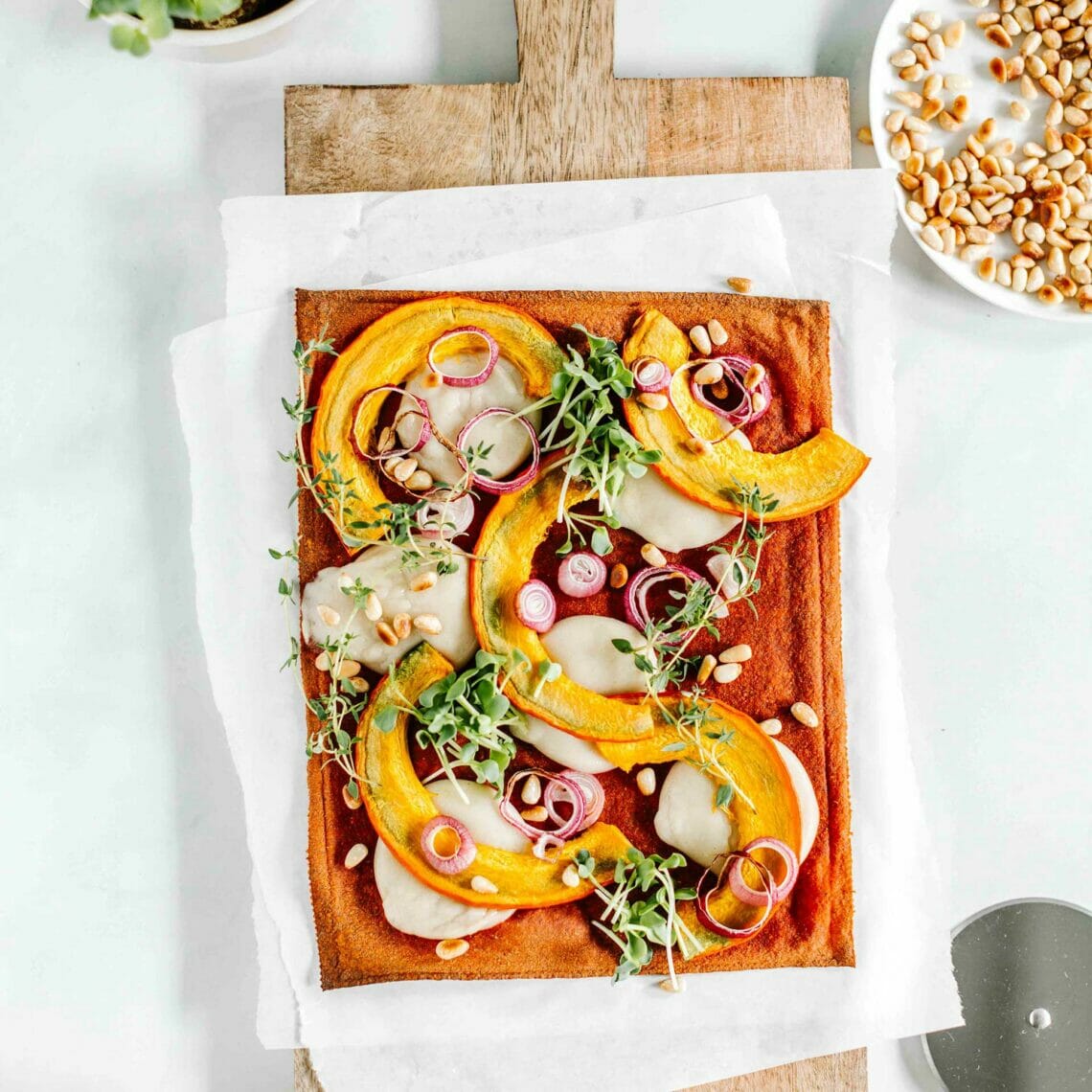 Pizzaboden auf Holzbrett belegt mit Mozzarella, Kürbis, Zwiebeln, gerösteten Pinienkernen, Sprossen und Thymian